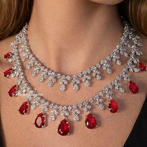Op de afbeelding: Twee-rijige ketting met een bloemmotief, bezet met heldere stenen en levendige rode, druppelvormige edelstenen. De sieraden worden gepresenteerd tegen een donkere achtergrond, waardoor de schittering van de edelstenen wordt benadrukt.
