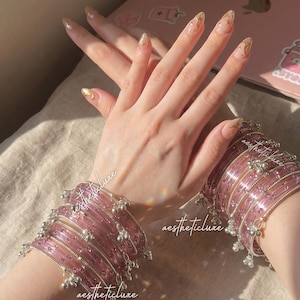 May include: Two sets of pink bangles with silver accents and small dangling charms. The bangles are stacked on the wrists, and the image is well-lit. The nails are painted with gold glitter. The text "aestheticticluxe" is visible.