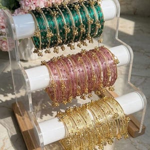 Puede incluir: Un surtido de pulseras en verde, rosa y dorado exhibidas en un soporte de acrílico transparente. Cada pulsera presenta un diseño delicado con pequeños dijes dorados colgantes. Las pulseras están dispuestas en filas ordenadas, mostrando sus intrincados detalles.