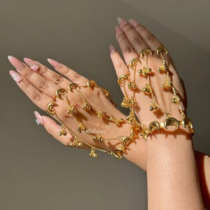 Könnte beinhalten: Goldfarbener Handschmuck mit Halbmond- und Glockenanhängern. Der Schmuck verbindet Ringe mit einem Armband durch zarte Ketten. Das Design umfasst mehrere Stränge kleiner, baumelnder Glocken.