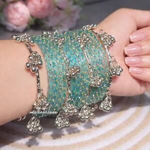 Puede incluir: Un conjunto de pulseras turquesas con detalles plateados y colgantes. Las pulseras están adornadas con pequeños adornos brillantes. Las joyas se exhiben en una muñeca, mostrando el intrincado diseño y la artesanía.