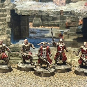 Puede incluir: Cinco figuras de fantasía en miniatura, cada una sobre una base redonda, posan frente a una estructura de piedra. Las figuras visten armaduras y ropa roja y plateada, y sostienen armas como espadas y lanzas. Las figuras tienen una apariencia amenazante.