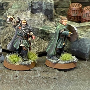 Könnte beinhalten: Zwei Miniatur-Fantasy-Figuren, eine mit einer Laterne und die andere mit einem Schild. Beide Figuren tragen grüne Umhänge und stehen auf einem felsigen Gelände. Die Figuren sind detailliert bemalt.