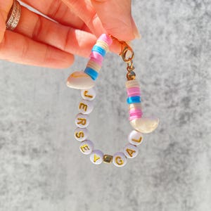 May include: A white beaded bracelet with the name "JERSEY GAL" spelled out in gold letters. The beads are interspersed with pink, blue, and white striped beads.