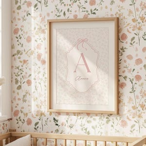 Op de afbeelding: Een ingelijste print met de roze letter "A" en de naam "Annie" op een patroonachtergrond. De print zit in een licht houten frame, tegen een bloemenbehang. Een wieg met kussens staat op de voorgrond.