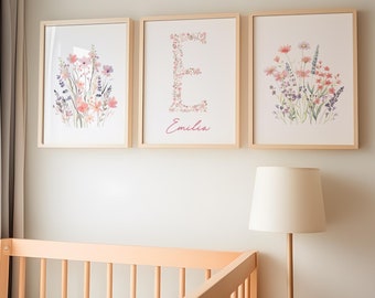 May include: Three framed prints with watercolor floral designs. The center print features the letter "E" made of flowers and the name "Emilia" written below. The prints are hung on a white wall above a wooden crib.