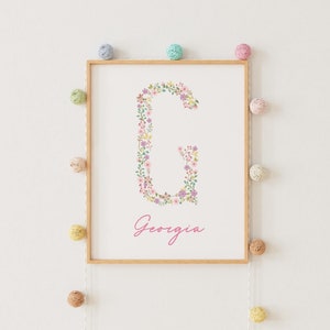 Può includere: Una stampa incorniciata con una lettera G floreale e il nome Georgia scritto in corsivo rosa sotto. La stampa è circondata da una cornice in legno chiaro ed è appesa a una parete bianca.