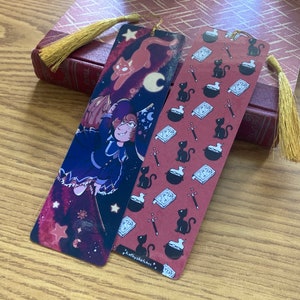May include: Two bookmarks with a witch and a cat design. The first bookmark features a witch flying through the night sky with a cat and stars. The second bookmark has a repeating pattern of black cats, cauldrons, and wands on a red background. Both bookmarks have gold tassels.