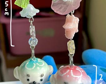 Puede incluir: Dos llaveros con dijes que representan figuras de animales de cerámica blanca con detalles azules y rosas. Los dijes están unidos a cadenas plateadas con diversas cuentas y dijes, incluyendo flores, hojas y una perla.