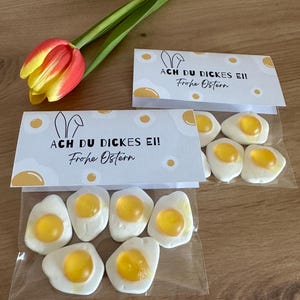 Könnte beinhalten: Zwei weiße Tüten mit einer weißen Karte, auf der "Ach du dickes Ei! Frohe Ostern" steht, und einer gelben Tulpe. Die Tüten enthalten gelbe und weiße Gummibärchen in Form von Spiegeleiern.