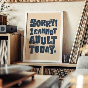Peut inclure: Une affiche encadrée avec le texte "SORRY! I CANNOT ADULT TODAY." en lettres bleu foncé. L'affiche est dans un cadre blanc et repose sur une étagère en bois. L'arrière-plan comprend un tourne-disque, des disques et des étagères en bois.