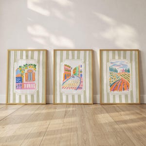 Op de afbeelding: Drie ingelijste kunstdrukken met kleurrijke illustraties van Italiaanse scènes. Elke print heeft een gouden lijst en een gestreepte achtergrond. De kunstwerken tonen een balkon met citroenen, een geplaveide straat en een landelijk landschap.