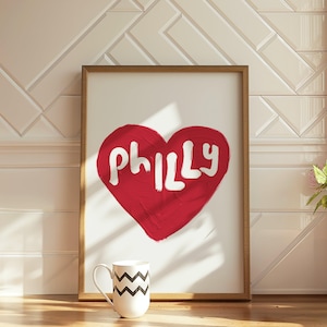 Könnte beinhalten: Ein gerahmter Kunstdruck mit einem roten Herz und dem Wort "Philly" in Weiß. Vor dem Kunstwerk steht eine weiße Tasse mit einem schwarzen Zickzackmuster. Der Rahmen ist hellbraun.