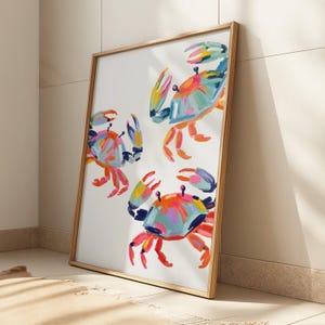 May include: A framed art print featuring three colorful crab illustrations. The crabs are painted with vibrant hues of blue, orange, pink, and yellow against a white background. The artwork is encased in a natural wood frame, adding a touch of coastal charm.
