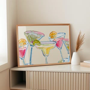Op de afbeelding: Een ingelijste kunstdruk met een aquarel schilderij van verschillende margarita cocktails. De drankjes zijn in verschillende kleuren, waaronder roze, geel en groen, en gegarneerd met limoen- en sinaasappelschijfjes. De lijst heeft een lichte houtkleur.