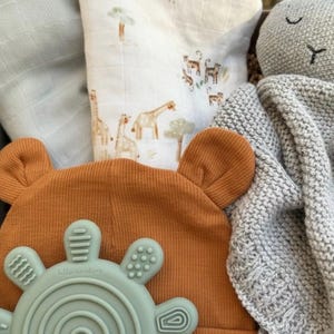May include: A baby gift set featuring a rust-colored beanie with bear ears, a mint green teether, a gray knit blanket, and a muslin swaddle with safari animal illustrations. The teether has the text "hello sunshine".