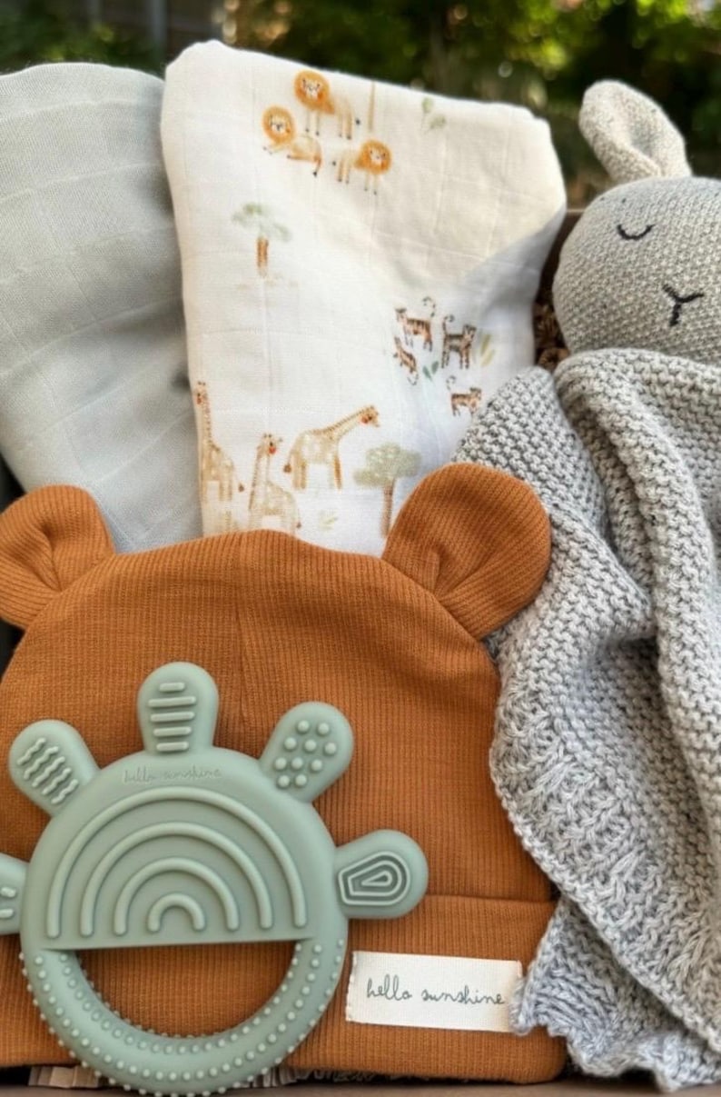 May include: A baby gift set featuring a rust-colored beanie with bear ears, a mint green teether, a gray knit blanket, and a muslin swaddle with safari animal illustrations. The teether has the text "hello sunshine".