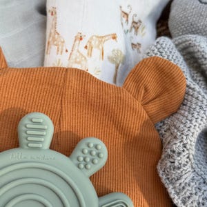 May include: An orange baby hat with bear ears, a blue-green teething toy with a rainbow design and the text "hello sunshine", and a white blanket with giraffe illustrations. A gray knitted blanket and a gray stuffed animal are also visible.