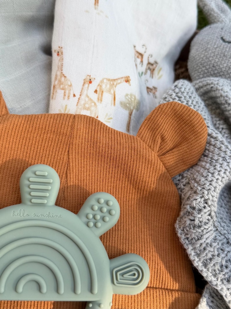 May include: An orange baby hat with bear ears, a blue-green teething toy with a rainbow design and the text "hello sunshine", and a white blanket with giraffe illustrations. A gray knitted blanket and a gray stuffed animal are also visible.