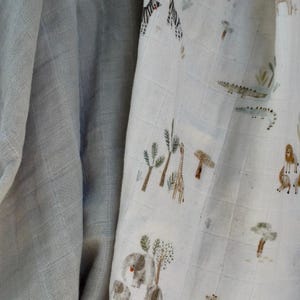 May include: A light blue muslin blanket with a safari animal print. The print features elephants, giraffes, zebras, and crocodiles in a naturalistic style. The blanket is soft and lightweight, ideal for a baby.