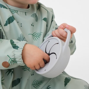 Puede incluir: Un niño sostiene una taza para snacks gris claro con asa. La taza tiene una abertura flexible. El niño lleva un babero verde claro con un estampado de hojas tropicales. El babero tiene mangas largas y cuello redondo.