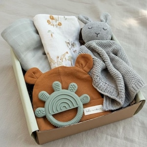 May include: A baby gift set in a cardboard box. The set includes a brown bear-eared hat, a green teether, a gray knitted blanket, a white printed swaddle, and a gray bunny plush toy. The swaddle has a safari animal print.