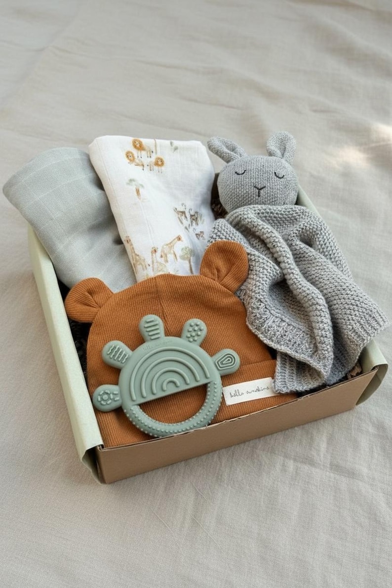 May include: A baby gift set in a cardboard box. The set includes a brown bear-eared hat, a green teether, a gray knitted blanket, a white printed swaddle, and a gray bunny plush toy. The swaddle has a safari animal print.