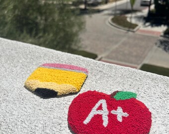 May include: A red apple with a white "A+" on it and a yellow pencil with a pink eraser are on a white surface.