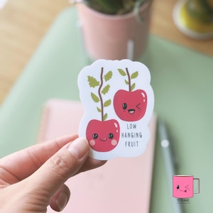 May include: A white sticker featuring two cartoon cherries with smiling faces and green stems. The text "LOW HANGING FRUIT" is below the cherries. The sticker is held by a hand.