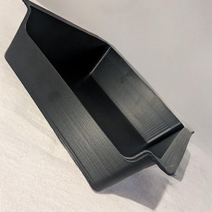 Puede incluir: Una bandeja de almacenamiento negra y rectangular con una superficie texturizada. La bandeja tiene un diseño angular único con bordes elevados y un divisor central. La bandeja está vacía y descansa sobre una superficie blanca.