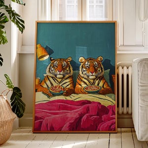 Puede incluir: Una impresión artística enmarcada que presenta a dos tigres en la cama, cada uno sosteniendo un cuenco de comida. La obra de arte tiene un fondo verde azulado, una manta rosa y un marco de madera. Se ve una pequeña lámpara naranja.