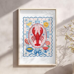 May include: Framed cross-stitch art featuring a red lobster in a pink oval, surrounded by blue and white patterns. Includes lemon slices, a seashell, and a blue bow. The artwork is in a light wooden frame.