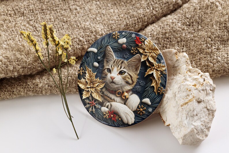 May include: A round decorative plate featuring a tabby cat with a festive collar and a bell. The plate has a dark blue background with gold poinsettias, snowflakes, and sprigs of evergreen. A sprig of dried yellow flowers is to the left.