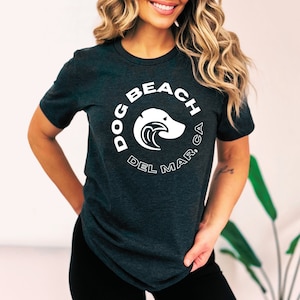 May include: A dark grey t-shirt with a white graphic of a dog head with a wave behind it. The text "DOG BEACH DEL MAR, CA" is written in a circle around the dog.