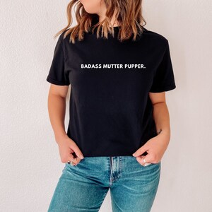 May include: A black t-shirt with white text that reads "BADASS MUTTER PUPPER."