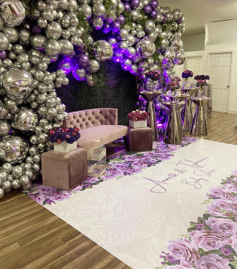 Puede incluir: Un tel&oacute;n de fondo blanco con globos morados y plateados, un sof&aacute; morado y un suelo con estampado floral con el texto "Annette & Typhonne Baby Shower" impreso en &eacute;l.