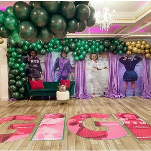 Puede incluir: Una sala de fiestas decorada con globos verdes y dorados, cortinas moradas y grandes letras rosas que deletrean "GIG" en el suelo. Las letras tienen im&aacute;genes de mujeres con diferentes atuendos y el texto "#HASTEMANHI" y "Hotter!" en ellas.
