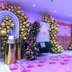 Puede incluir: Una fiesta de Sweet Sixteen con tem&aacute;tica morada y blanca con un gran arco de globos, un centro de mesa floral y un pastel de dos niveles. El tel&oacute;n de fondo presenta una pared de flores moradas y un letrero que dice "Sweet Sixteen".