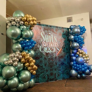 Puede incluir: Un tel&oacute;n de fondo para un evento decorado con globos azules, plateados y dorados. El tel&oacute;n de fondo tiene un patr&oacute;n floral azul verdoso y blanco y el texto "Suits + Sneakers + Cigars".