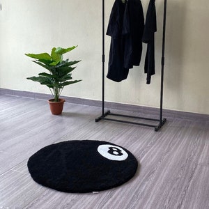 May include: A black and white 8-ball rug with a black metal clothing rack in the background. The rack has three black shirts hanging on it.