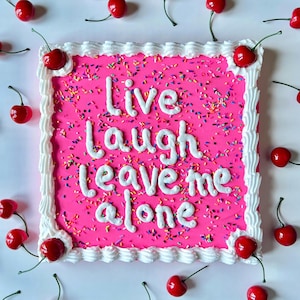 May include: A square pink cake with white frosting and the words "Live Laugh Leave Me Alone" in white icing. The cake is decorated with colorful sprinkles and surrounded by red cherries.