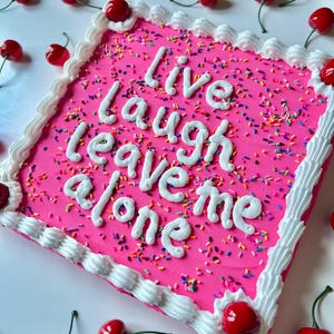 May include: A square pink cake decorated with white frosting, sprinkles, and red cherries. The cake has the words "Live Laugh Leave Me Alone" written in white frosting. Several red cherries with green stems are scattered around the cake.