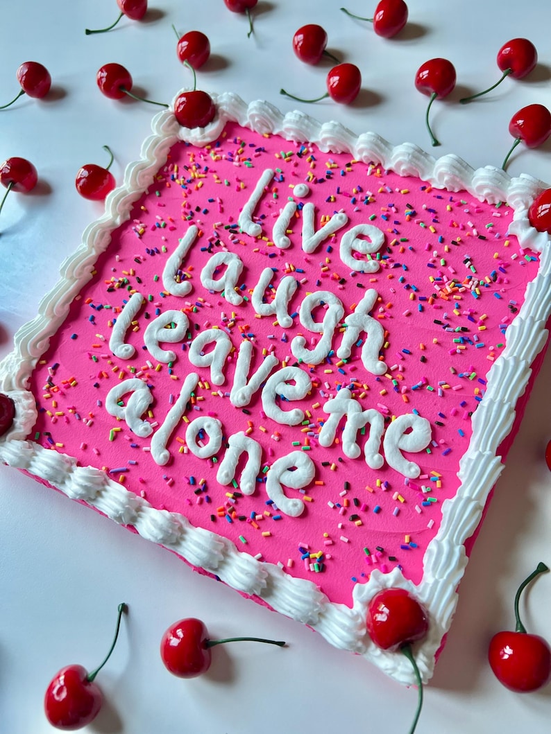 May include: A square pink cake decorated with white frosting, sprinkles, and red cherries. The cake has the words "Live Laugh Leave Me Alone" written in white frosting. Several red cherries with green stems are scattered around the cake.