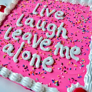 May include: A square pink cake decorated with white frosting, sprinkles, and cherries. The cake has the words "Live laugh leave me alone" written in white frosting. The edges are lined with white frosting.