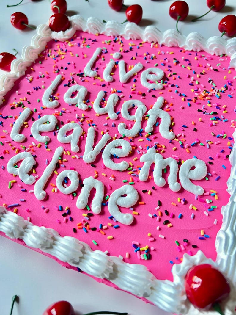 May include: A square pink cake decorated with white frosting, sprinkles, and cherries. The cake has the words "Live laugh leave me alone" written in white frosting. The edges are lined with white frosting.