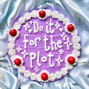 May include: A round, purple cake with white frosting and the phrase "Do it for the plot" written on top. The cake is decorated with white frosting swirls and red cherries.