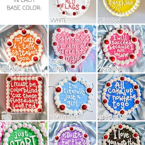 May include: A collection of decorated cakes with various base colors, including white, yellow, orange, light pink, hot pink, red, light blue, dark blue, green, purple, and black. Each cake features unique frosting and cherry decorations, with different phrases written on them.