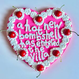 May include: A heart-shaped cake with bright pink icing, decorated with white piped frosting borders and seven red cherries. The text written in white icing reads: a hot new bombshell has entered the villa.