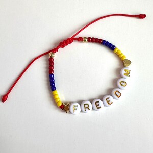 Peut inclure: Un bracelet en perles rouge, blanc et bleu avec le mot "FREEDOM" &eacute;crit en lettres dor&eacute;es sur des perles blanches. Le bracelet a une fermeture en cordon rouge.