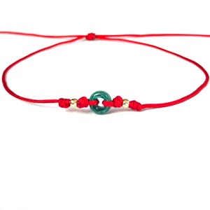 Puede incluir: Una pulsera de hilo rojo con una cuenta verde circular y dos peque&ntilde;as cuentas doradas. La pulsera est&aacute; anudada y es ajustable. El dise&ntilde;o es sencillo. El fondo es blanco.